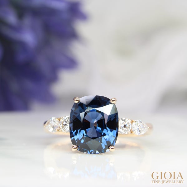 Blue Spinel Proposal Ring Featuring a unique bluish spinel elegantly set in 4-prongs, accentuated by round and pear-shaped cluster diamonds on each side.