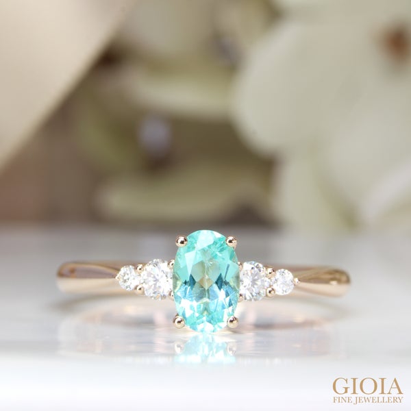 Paraiba Tourmaline Proposal Ring Featuring round cluster side diamonds embracing the Paraiba tourmaline, align perfectly with the sleek tapered knife edge bands in rose gold.