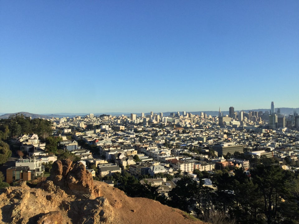 Clear blue sky over distant North East Bay hills in the distance, San Francisco’s tall buildings and downtown on the horizon, with more buildings nearer, trees from Buena Vista Park on the left, a rocky outrcopping from Corona Heights park immediately in the foreground.