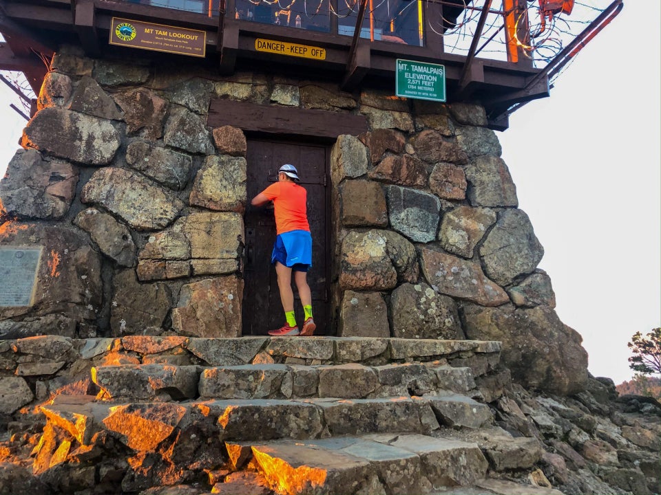Tantek touching the door of the rock and wood structure at the top of Mount Tam East peak, rock steps just below him, their left edges tinged in orange from the sunrise.
