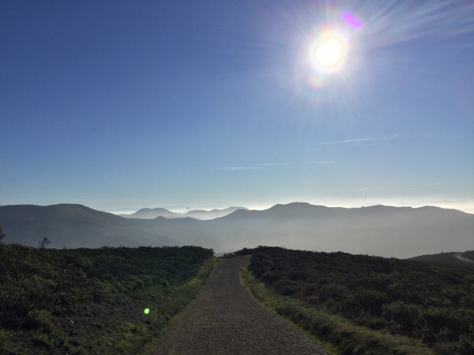 The sun shining above the Marin Headlands, low haze barely visible behind them, obscuring most of the tiny distant San Francisco skyline peaking through the hills, a wide trail in the foreground with low green bushes on either side.