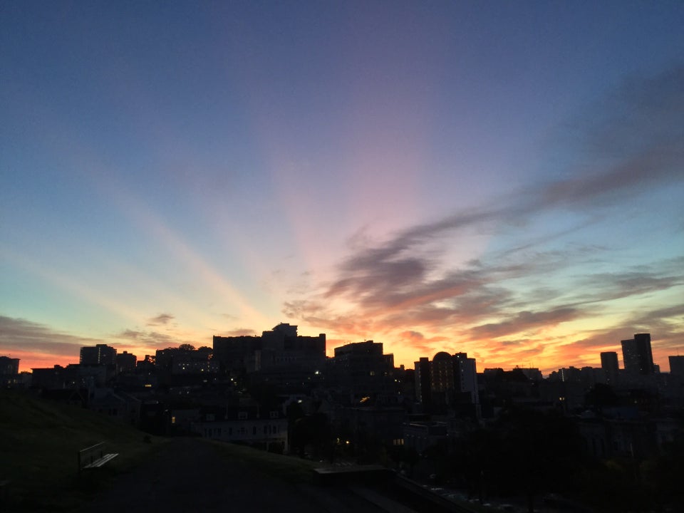 San Francisco cityscape backlit by orange sunbeams originating from near the center behind the buildings, against a blue sky with scattered clouds