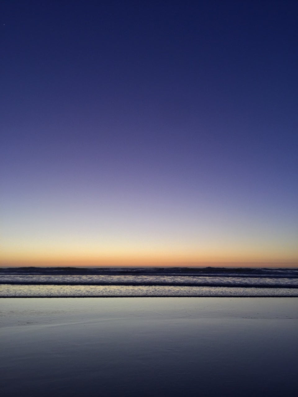 Deep blue sky with a gradient to purple then to orange at the horizon just above the ocean, gentle small waves, glistening sand reflecting light gray to darker blue gray.