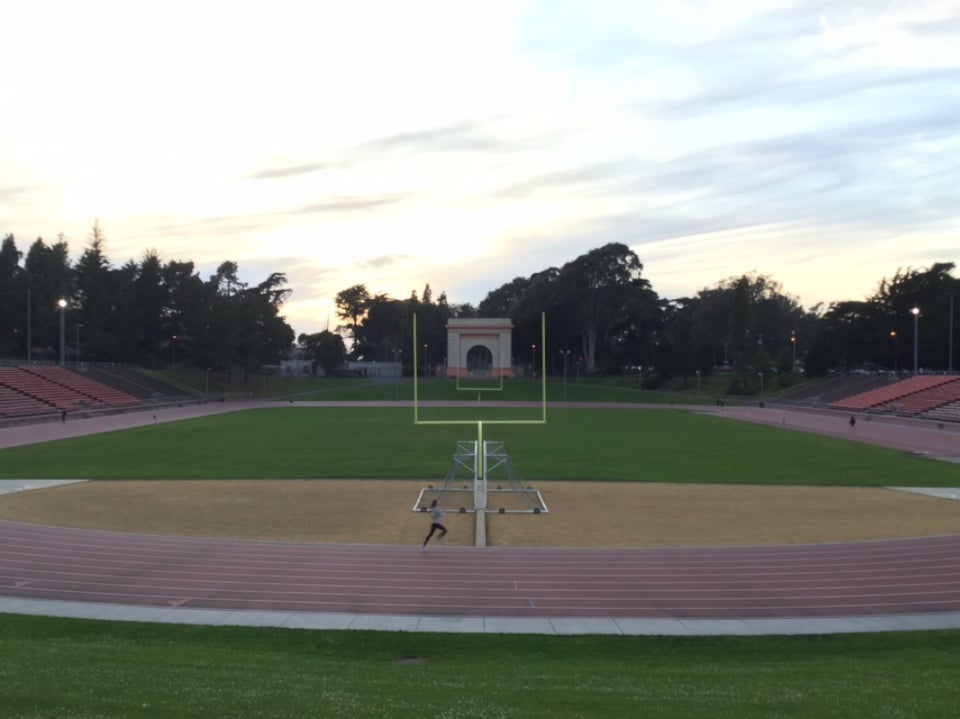 A bright sky overexposed by the setting sun lighting up clouds, trees in the distance above Kezar Stadium, football field goals aligned in the green field, a single woman runner sprinting in lane 1 on the track.