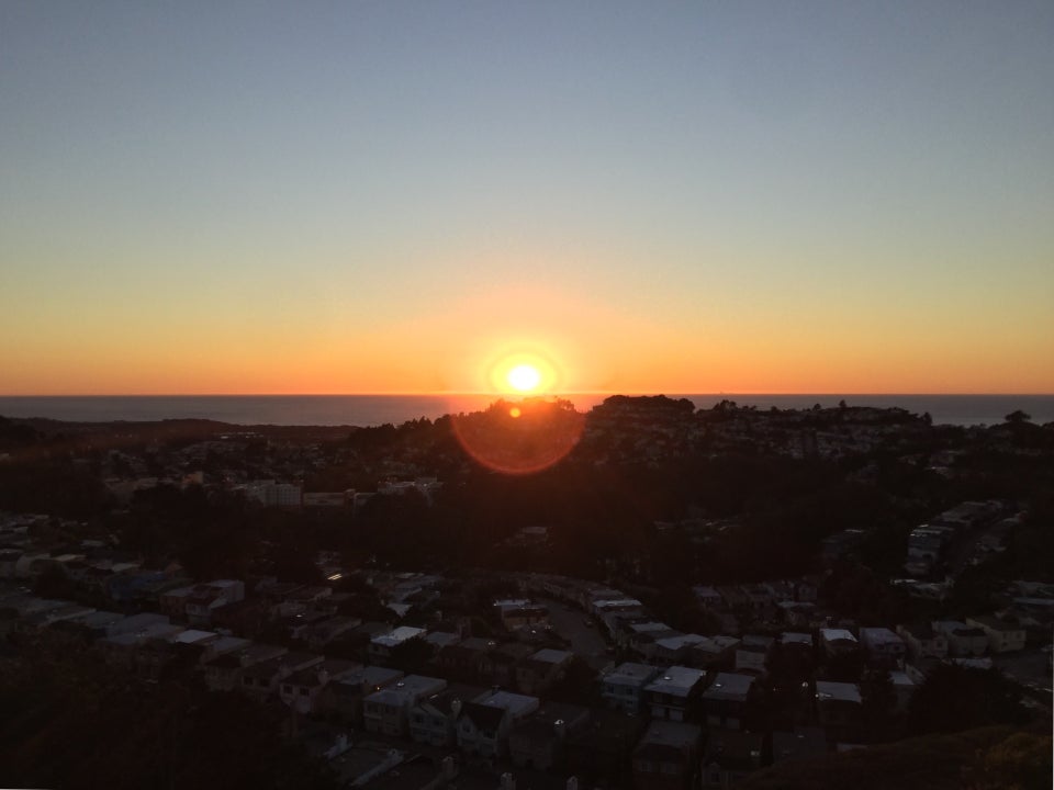 Sun setting over the Pacific Ocean, backlit dark trees on hills in the distance below, scattered houses on the hills in the shadow.