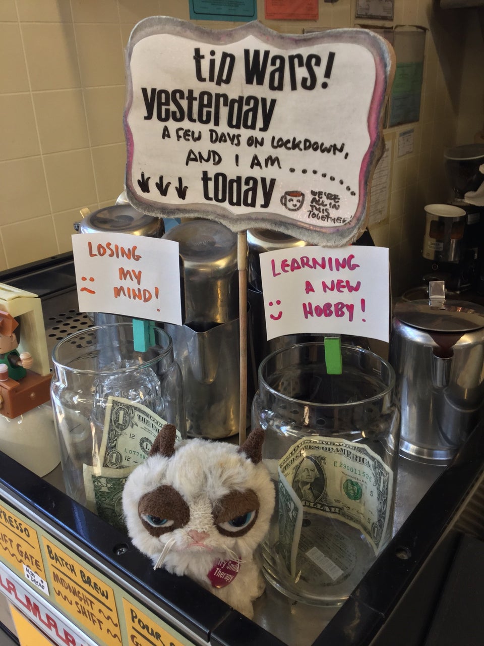 Tipping sign and two jars, one labeled Losing My Mind and the other Learning A New Hobby, each with a dollar inside, with a small Grumpy Cat stuffed doll in front of them wearing a small red pin saying Try Smile Therapy, all sitting on top of a professional espresso machine inside Haight Street Market.