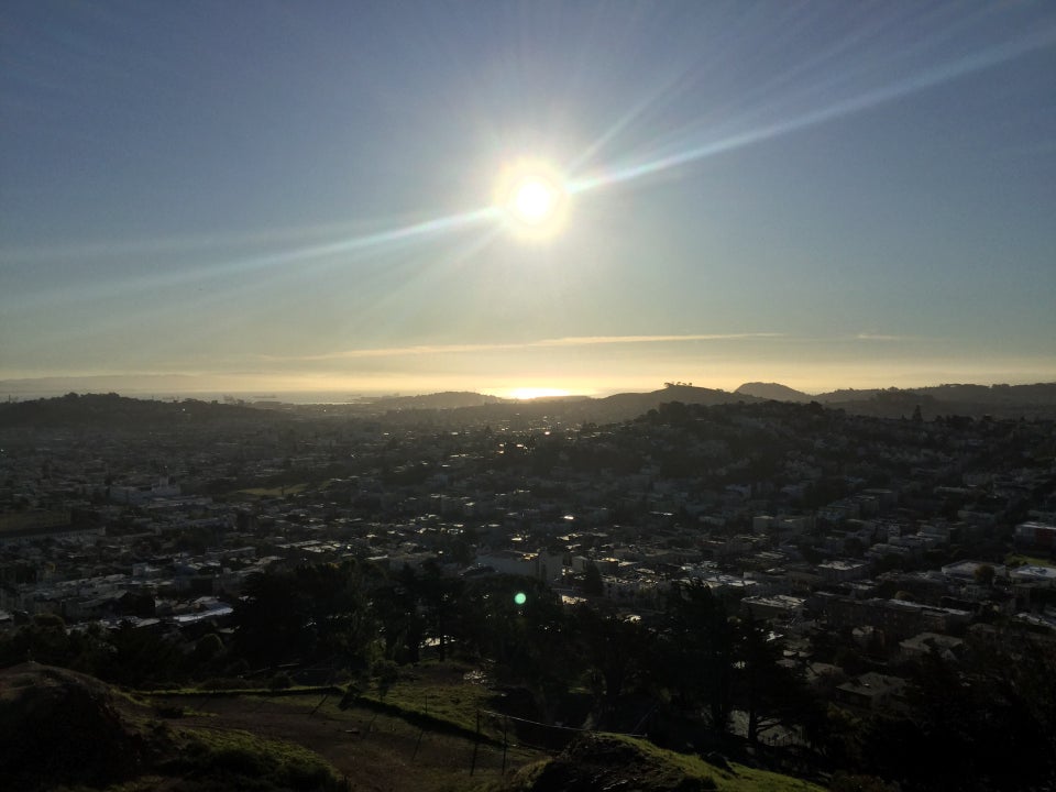 Sun over the bay, with sunbeams extending to the sides, and a sunlit bay, East Bay shoreline partially hidden by haze, San Francisco buildings in the middle, and the base of Corona Heights park below.