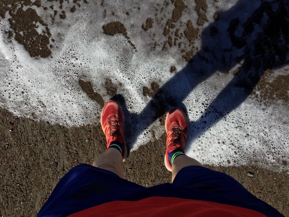 Looking down, standing on a coarse sandy beach with ocean surf lapping red trail shoes