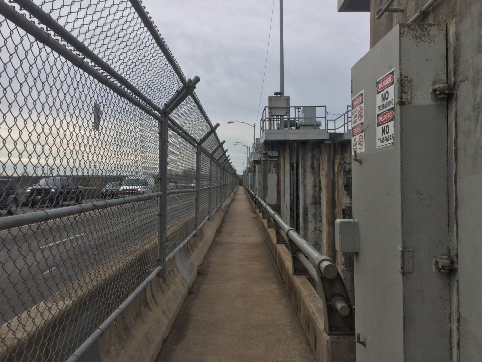 On top of the Longhorn dam, chainlink fencing on the left with the roadway on the other side, and the dam structures and warning signs on the right, the path across the river in between them.