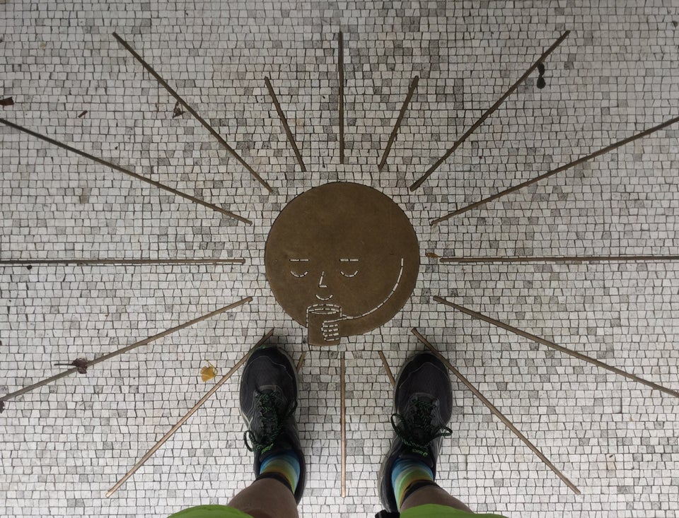Looking down at a mottled white mosaic entryway with a brass sun-with-a-face seemingly sipping a cup of coffee, brass segment sunbeams radiating outward.