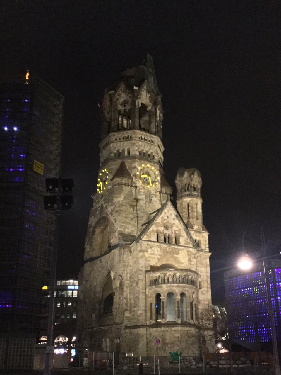 A old church with a severely damaged spire at night, with a light-up clock on it, with modern buildings with lights on both sides.