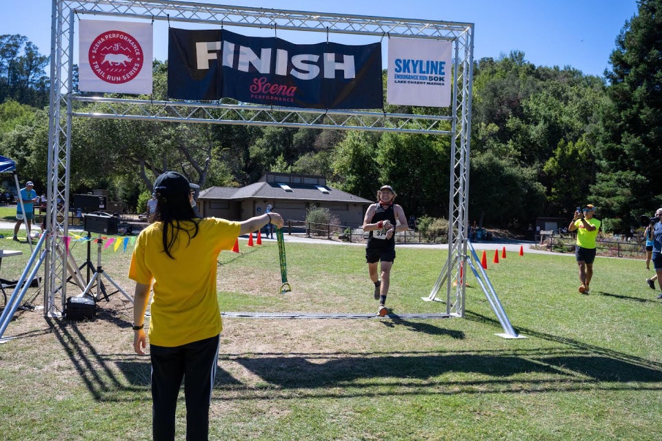 Tantek crossing the finish line sensor under the FINISH arch in the bright sunshine, smiling & stopping his watch, a volunteer in the foreground holding out a finisher medal, David Tran photographing from the side