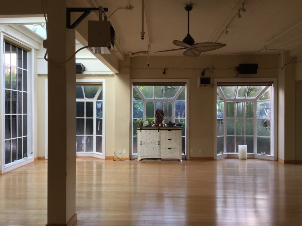 Yoga studio workout room with a heater and fan on the ceiling, a single support post on the left, steamed up multipaned bay windows across most of the walls letting in light and trees visible just beyond them, a small wide medium height cabinet set back in the center bay window with an alter to a small Ganesha statue on top, surrounded by small candles.