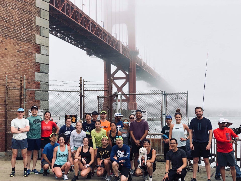 Runners lined up standing and kneeling in front of the Hoppers Hands fence with the Golden Gate Bridge in the background disappearing into the fog