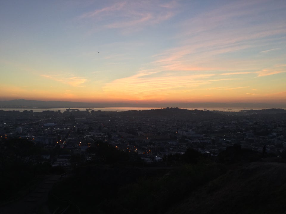 Pink and orange wisps of cotton candy clouds in the light blue pre-dawn sky above a horizon glowing yellow and orange over East Bay Hills, a sliver of bay visibe below it, then distant San Francisco hills, buildings with lights, and nearby hills and trails in darkness.