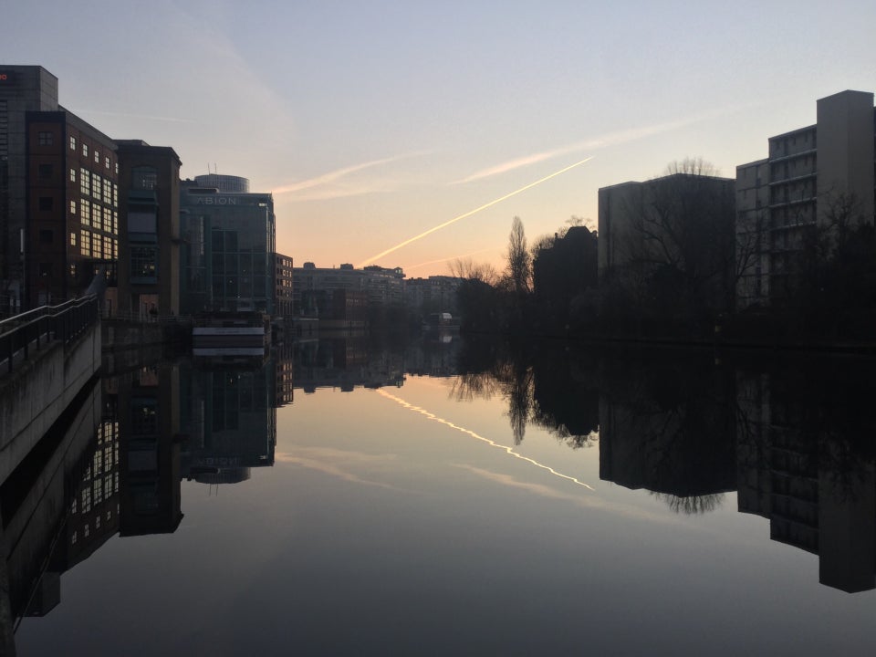 Clear sky with one contrail, slight orange glow towards the middle horizon, above buildings on both sides, trees on the right side of the river Spree which is showing their reflections.