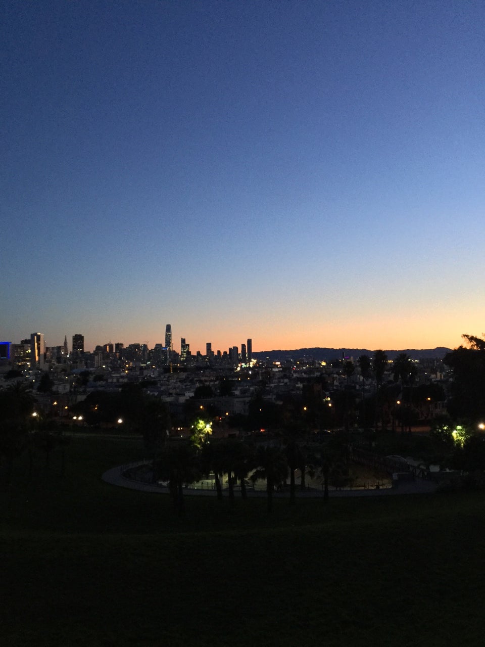 Clear blue to orange pre-dawn gradient sky above the San Francisco skyline in the distance behind a barely visible Dolores Park, a few paths and trees lit in by lamps.