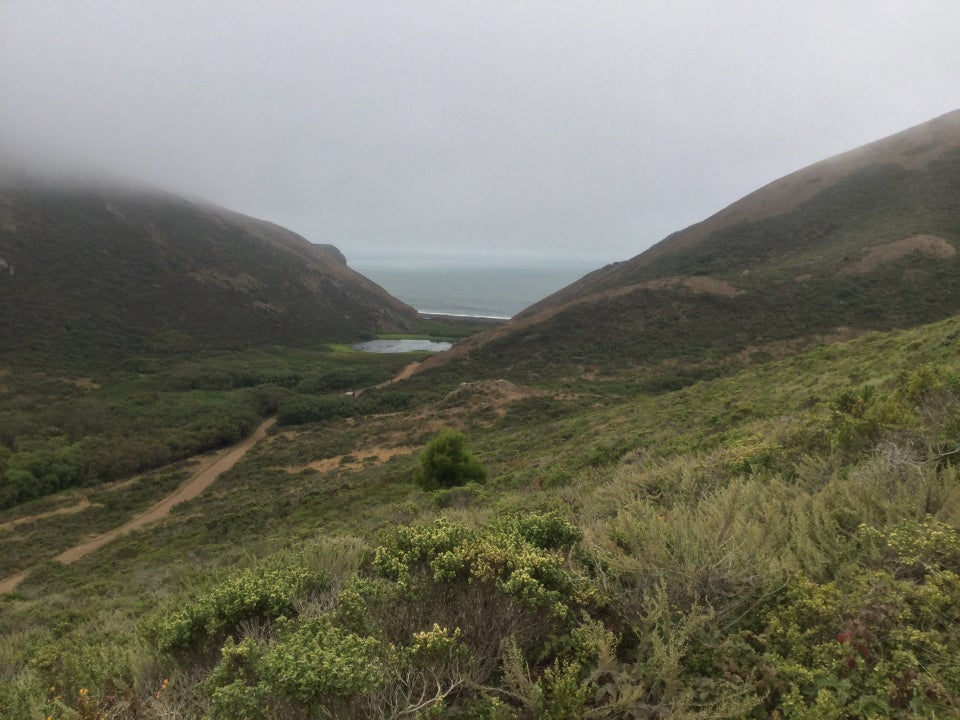 Tennessee Valley as seen from Coastal Fire road descent: green hills, Tennessee Valley trail winding them past a lagoon to the beach between two cloud covered hilltops.