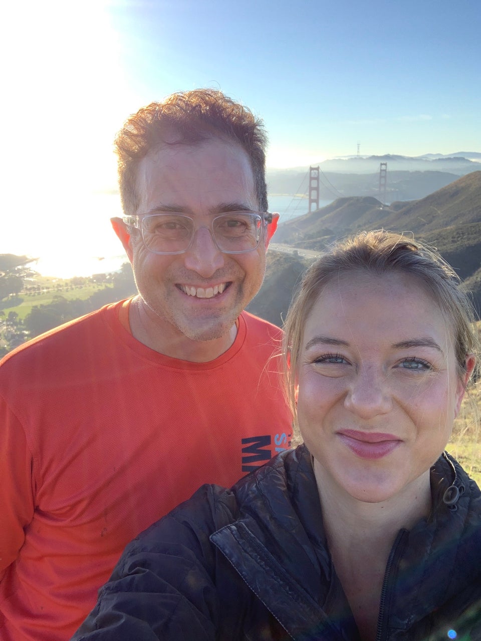 Emily taking a selfie of herself and Tantek with bright sun behind them, and the Golden Gate bridge towers poking above the Marin Headlands.