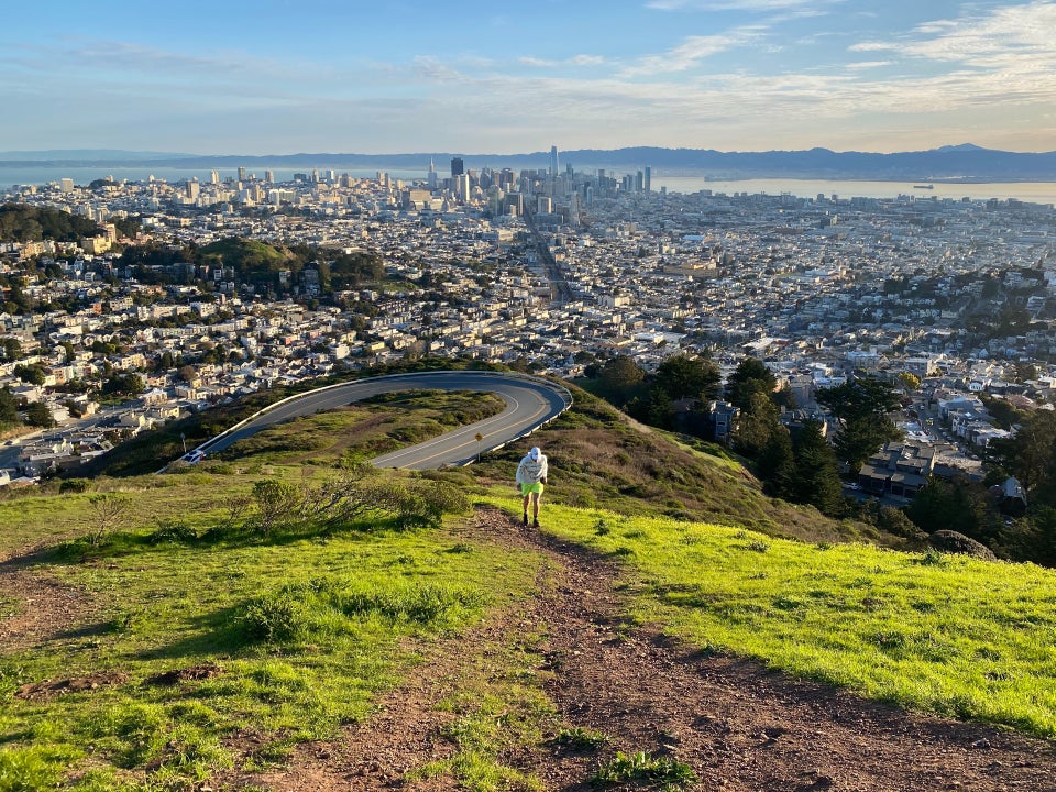 Clear morning sky with a few hazy clouds, East Bay hills behind a bay with a couple of tiny giant container ships, downtown San Francisco skyline and buildings, hairpin turn up Twin Peaks boulevard, light green grasses lit up with dew and morning sun, Tantek running up the trail between them to the top of Twin Peaks.