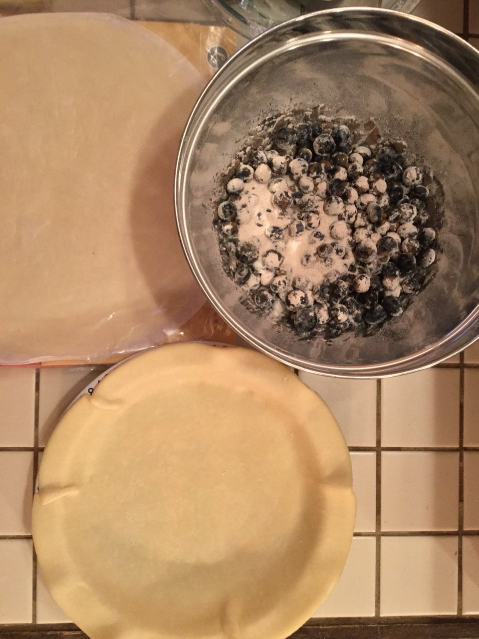 Blueberries and pie ingredients mixed in a metal bowl, pie crust lining a pie dish, atop an off-white tile countertop.