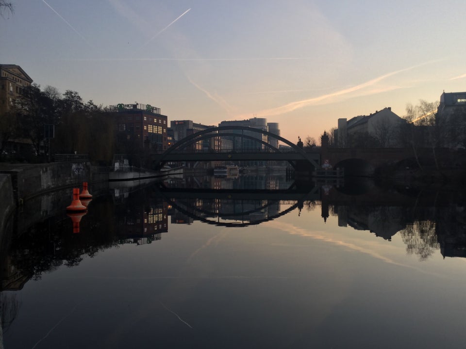 Clear sky with one contrail, slight orange glow towards the middle horizon, above buildings on either side of the river Spree which is showing their reflections.