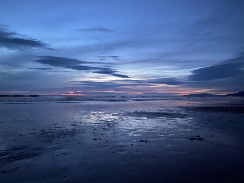 Medium blue to light blue post-sunset skies with ominous clouds and dark clouds on the horizon, with only slivers of orange from the sun, over crashing waves and a wet but rough shore, providing a distorted reflection of the sky and clouds above.