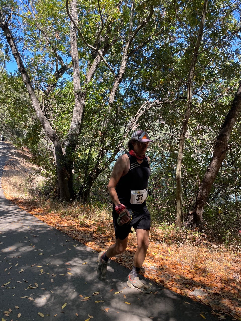 Tantek fast-walking downhill on an asphalt path in the shade next to some dry grass and trees, a lake peaking thru between their branches.