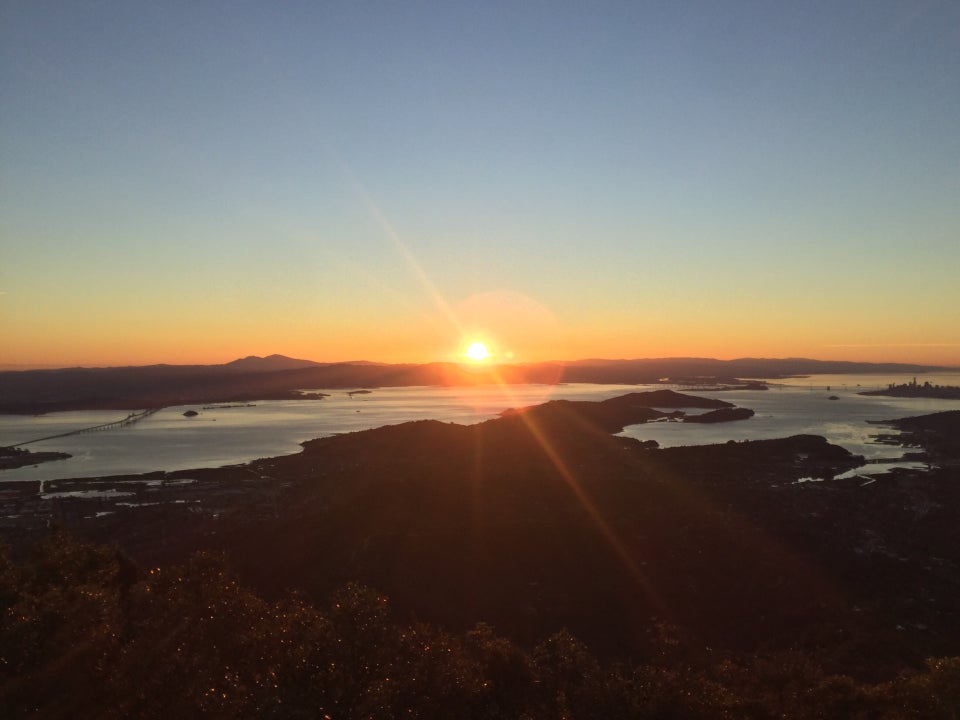 Clear blue to yellow and orange sky with the sun freshly rising with sunbeams in the middle, having just crested East Bay hills, the distant Mount Diablo a bit to the left, East Bay hill still in the dark, a tiny Richmond Bridge, the bay slowly lighting up from the sun, tiny Bay Bridge and San Francisco downtown skyline, Tiburon Peninsula and the nearby hills also still in the dark.