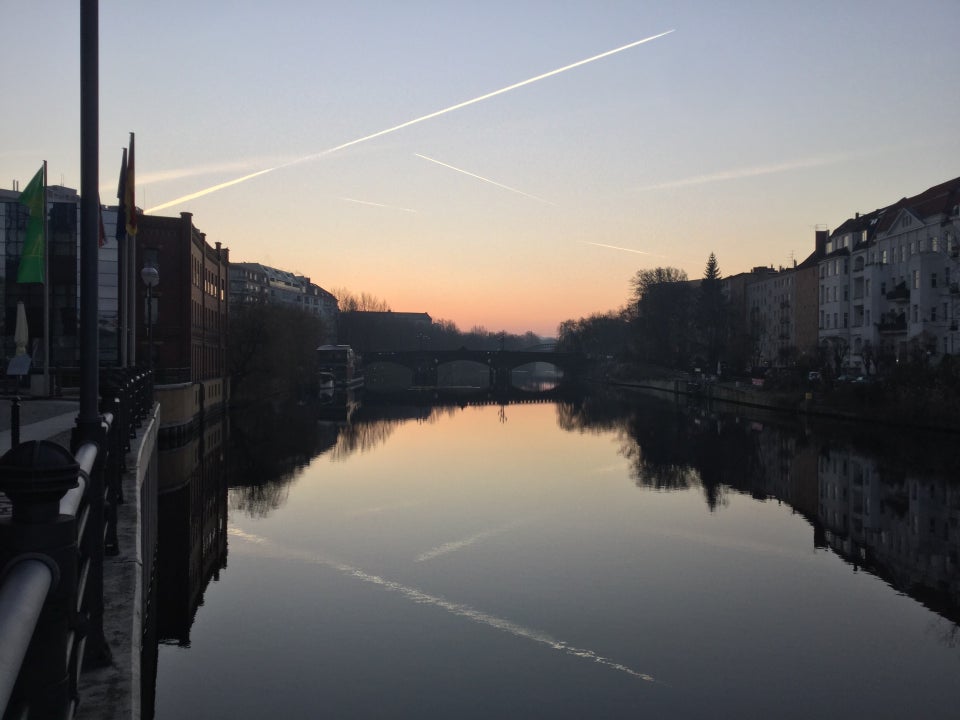 Clear sky with several contrails, orange glow in the middle of the horizon, above buildings on both sides, trees on the right side of the river Spree which is showing their reflections.