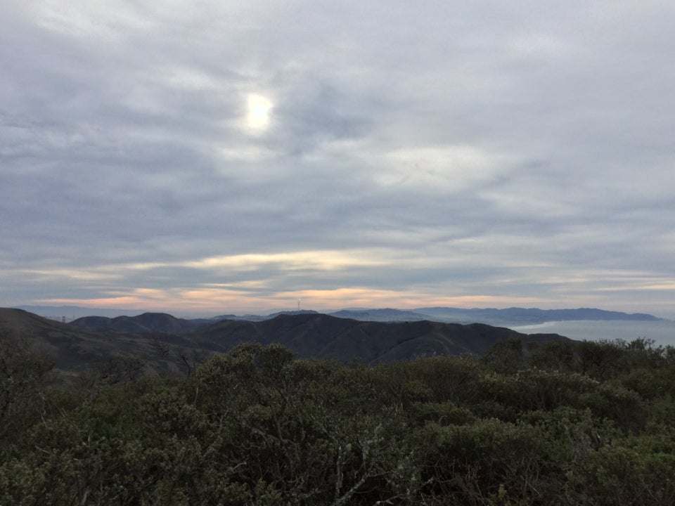 The sun shining through grey cloud cover, above a streaky orange horizon, hills of San Francisco Marin, the Pacific ocean on the right edge betwen them, green hills and buses in the foreground.