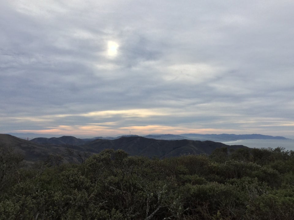 The sun shining through grey cloud cover, above a streaky orange horizon, hills of San Francisco Marin, the Pacific ocean on the right edge betwen them, green hills and buses in the foreground.