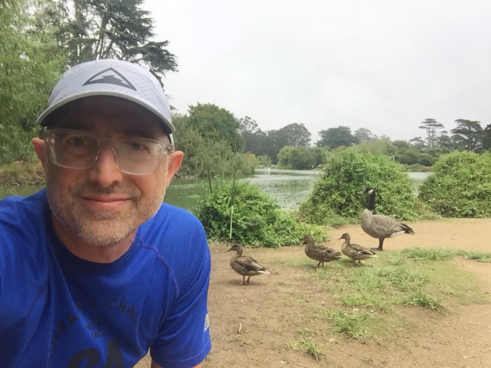 Tantek taking a selfie with three ducks and a goose behind him on the short of Stow Lake, bushes and green lake water behind them, part of the island and trees on the opposite shore behind that under an overcast sky.