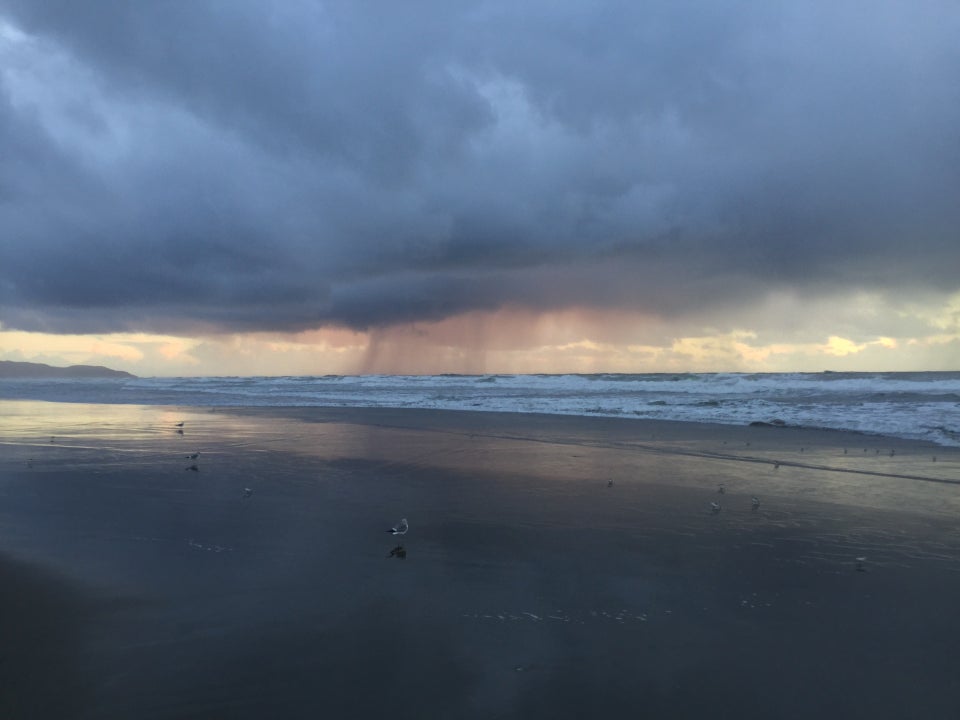 Thick clouds above a thin band of orange horizon backlighting some strands of rain falling from the clouds to the ocean, behind small crashing waves and a wet smooth sandy beach, a few birds on the sand looking for eats.