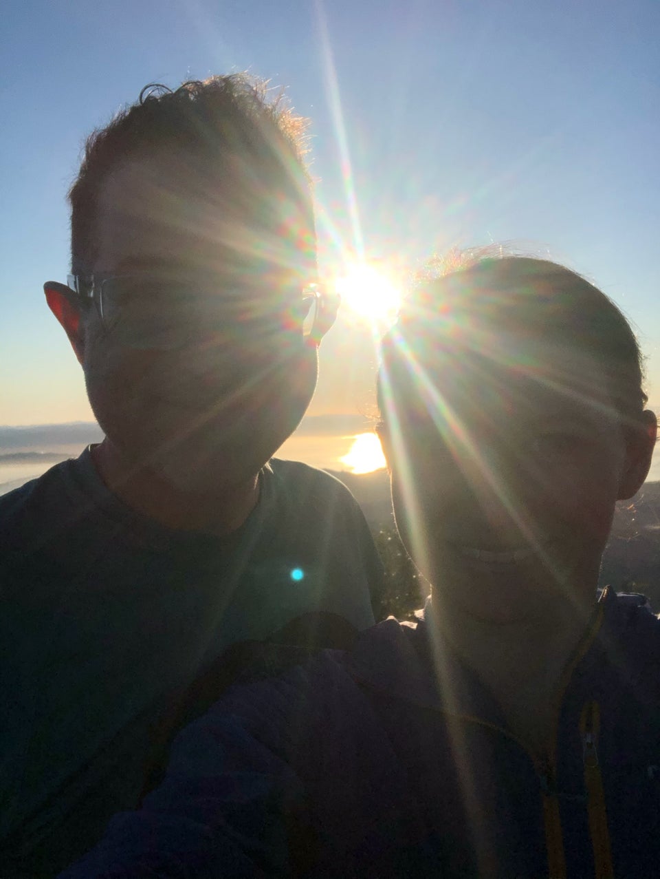 Tantek and Hannah with the sun shining brightly between them, sunbeams across their faces, the bay in the distance glowing with the sun, tiny East Bay hills in the distance.