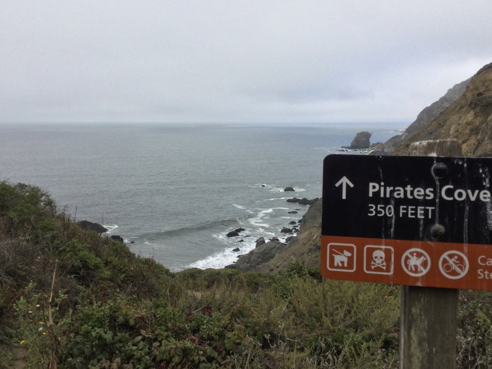 Looking down on Pirates Cove beach and across the Pacific Ocean, diffuse white light from the clouds, with a sign in the foreground indicating 350 feet to the cove, with various symbols.