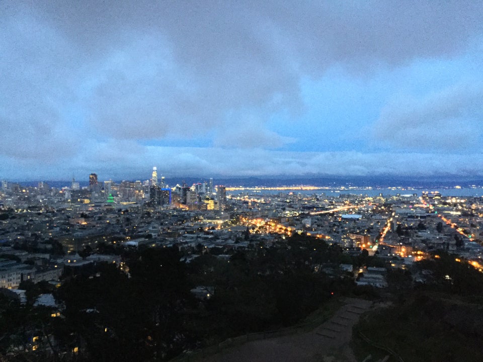 Grey clouds and bits of blue sky above white clouds covering the horizon in the distance above East Bay hills at dusk, downtown San Francisco buildings already lit, City Hall in green, buildings and streets lined with lights, trees below in darkness on the edge of barely visible beige gray trails and dirt area at Corona Heights park below.