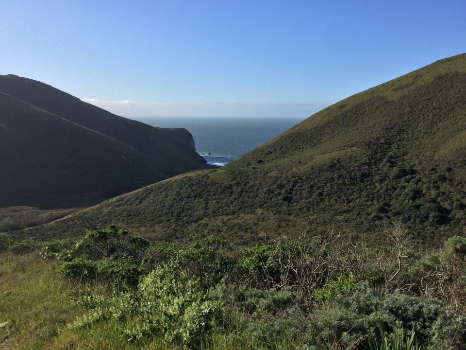 Clear blue sky above the green hills on either side of Tennessee Valley in Marin, where a bit of ocean is visible between them, as viewed downward from the Coastal Fire Road.