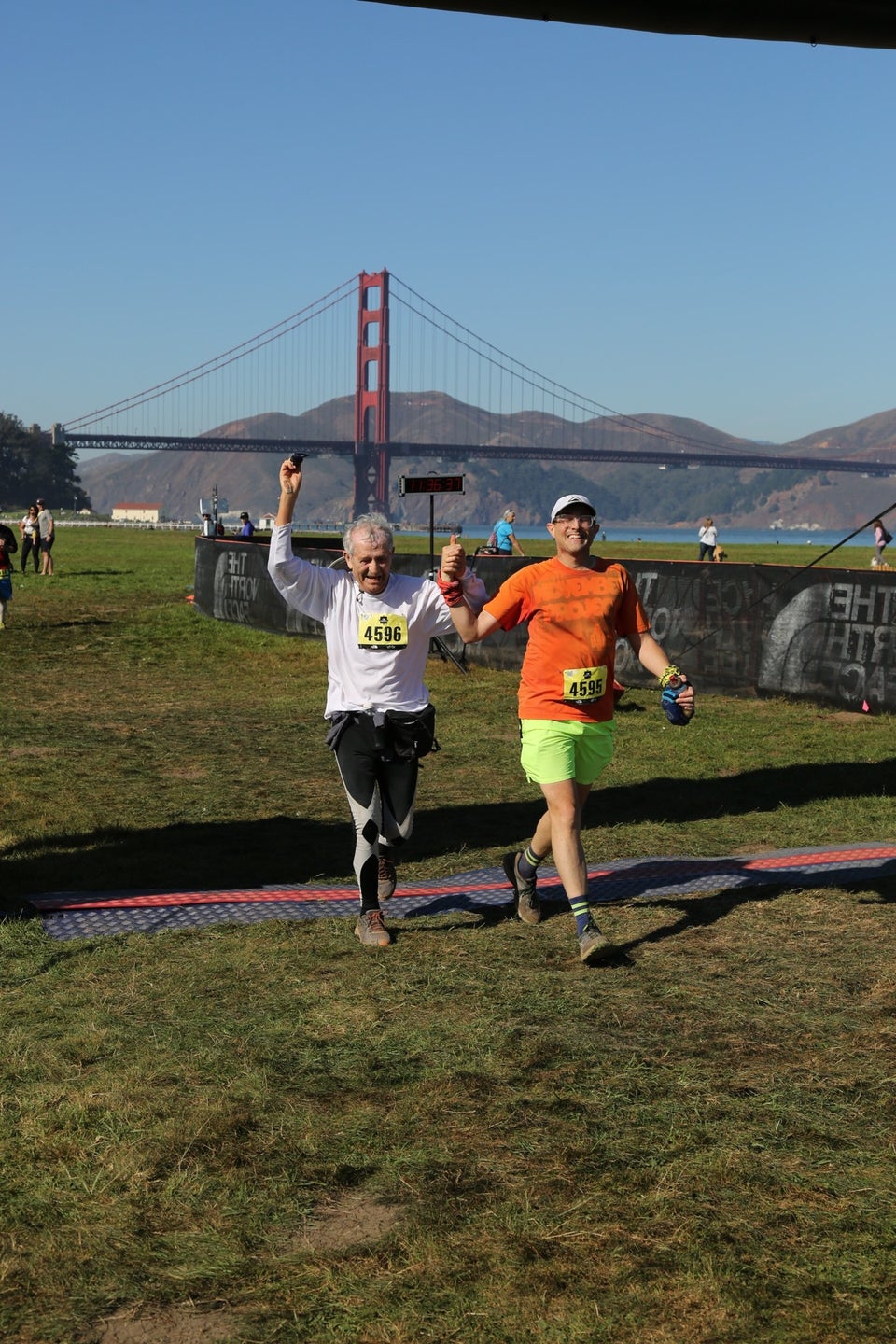 Hasan and Tantek smiling side by side holding raised hands between them, just after they crossed The North Face Endurance Challenge finish line in Crissy Field, San Francisco, with the Golden Gate Bridge and Marin Headlands in the background under a clear blue sky.