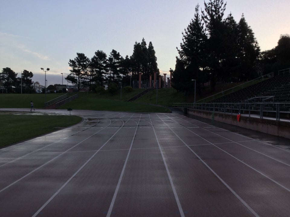 Light blue dawn sky above backlit trees just above the entrance to Kezar Track, as viewed from behind the starting area on the track, looking forward, and the somewhat wet track curving to the left.