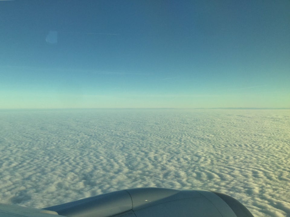 Clear blue sky above a hazy horizon and a blanket of clouds, the top of a jet engine just visible in the lower sixth of the photo.