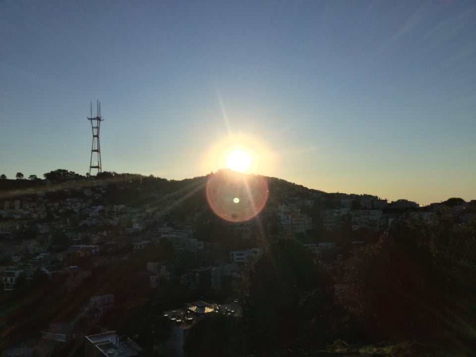 Clear blue sky with Sutro tower on the left and the sun setting behind a hill with sunbeams and a promient disc lens flare just below it, houses in the shadow on the hill below.