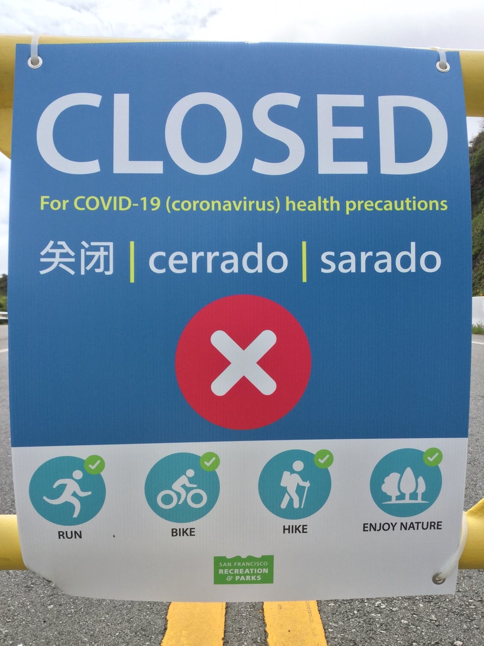 Close-up of a sign stating CLOSED For COVID-19 (coronavirus) health precautions, translations in Chinese & Spanish, a big red circle with a white x in the middle, and below that a row of four icons of activities with checkmarks (white on green background) in their top right corners, for run, bike, hike, and enjoy nature, below those a small San Francisco Recreation & Parks logo.