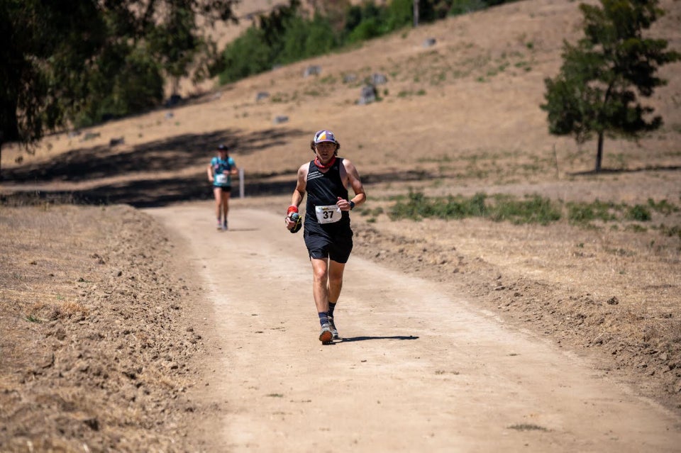 Tantek running on a wide smooth dirt trail with plowed rough dirt edges, bits of grass and a tree in the background nearby with many more trees in the distance, a single runner out of focus about 50 meters behind hime.