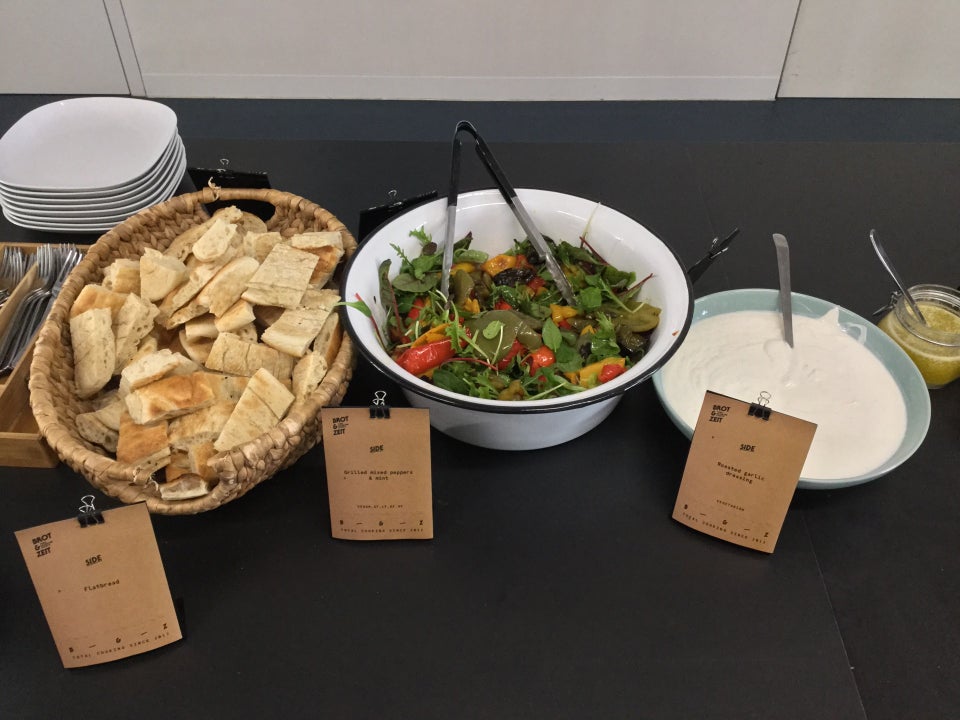 Long black table in front of a white wall, with plates, silverware, and catered food serving dishes of flatbread, salad, and two kinds of dressing.