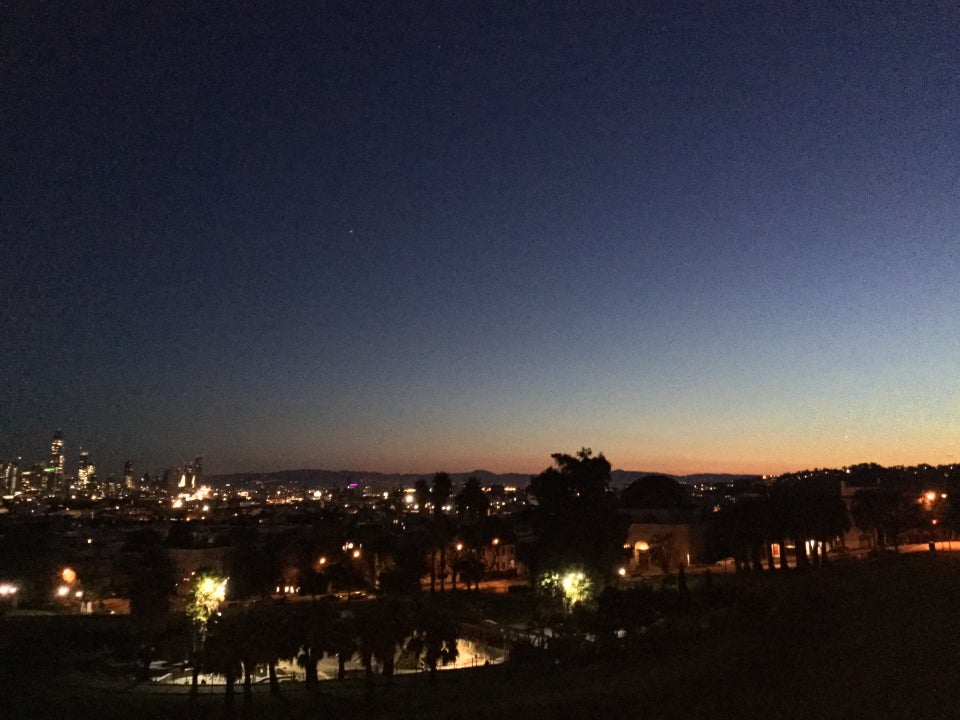 Dark blue sky above the San Francisco skyline, buildings lit, an orange yellow glow most pronounced towards the East, dark Dolores Park in the foreground, dotted with street lamps.