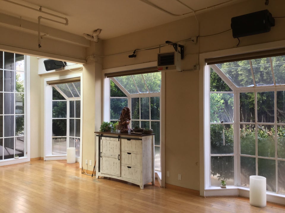 Inside of a yoga studio with hardwood floors, several wide floor to person height windows, each subdivided into a rectangular grid, three of them pushed outward in the style of bay windows, a medium sized distressed wood painted white dresser with a hardwood top holding a plant, candles, and a small Ganesha statue, above ceiling with several pipes running along the top painted the same color as the ceiling, a few room heaters mounted well above person height.