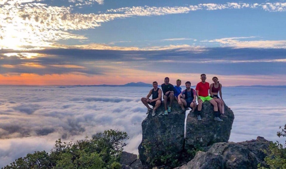 Trail runners perched on a rocky outcropping on top of Mt Tam, above the clouds, Mt Diablo in the background, the sun rising through the clouds