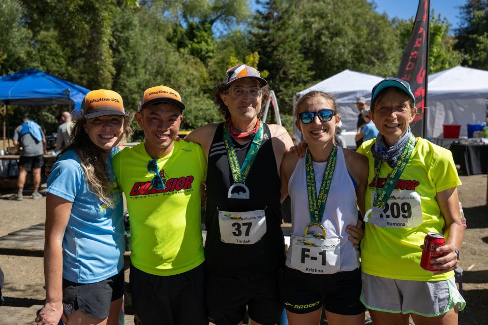 Trail runners Amanda, David Tran, Tantek, Kristina, Ramona standing side-by-side with arms around each other smiling, in front of the race finish area, the latter three still wearing their bibs and finish medals.