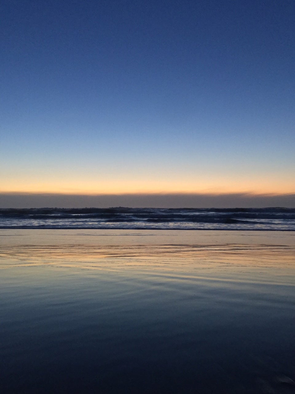Medium to light blue gradient of sky above an orange glowing horizon over low clouds, waves crashing below them, wet sand reflecting the orange glow and blue sky.